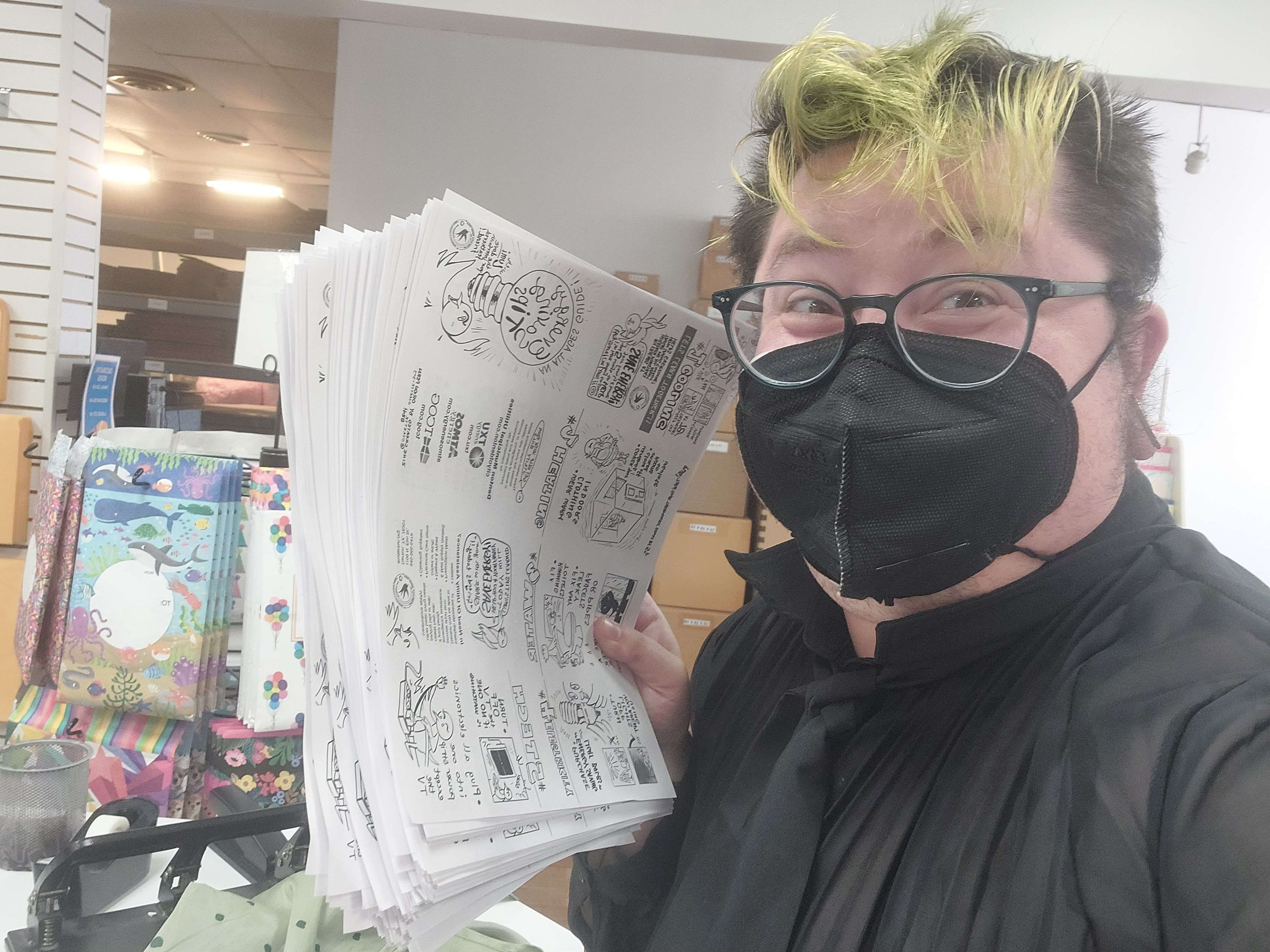 Me holding 250 zines for Interfaith Minstries of Denton, it is a thick stack of paper. I am a white trans man wearing glasses and a black N95 respirator mask. I am also wearing a black button up sheer shirt. I am in a print shop. I am smiling from under my mask.
